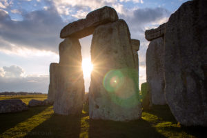 arch made of three large rectangular rocks at stonehenge with the setting sun appearing between them creating lens flare on the photograph