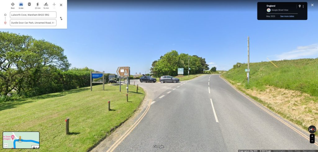 screenshot of Google Street View showing a tarmac country road between two grassy banks. there is a turning on the left with a brown arrow shaped road sign for Durdle Door pointing left.