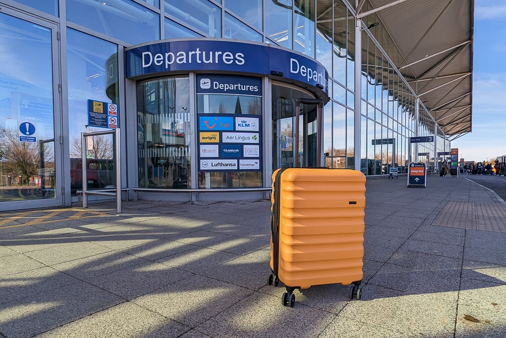 yellow hard suitcase on the pavement outside a revolving door labelled departures in the glass exterior wall of bristol airport