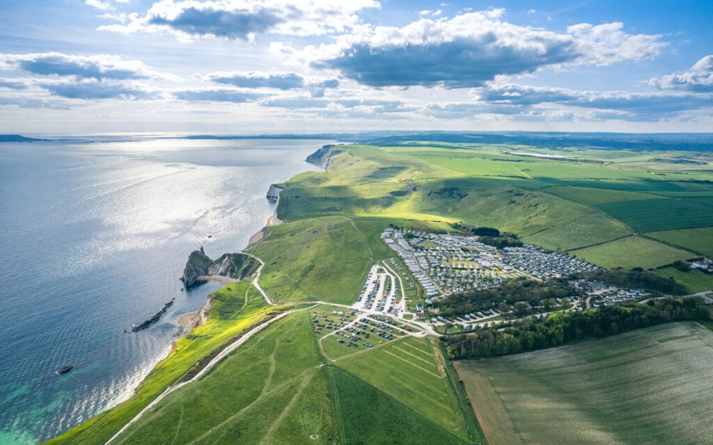 aeiral view of a green clifftop with a carpark and a holiday park filled with caravans in lines with the sea to the left on a sunny day. Durdle Door car park viewed from the air.