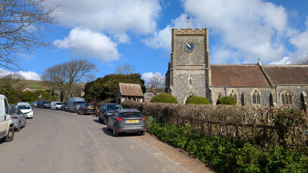 unmarked road in a village with cars parked on either side and a small stone church with a square clock tower to the right.