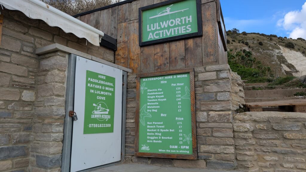 Stone wall with a green sign listing prices for kayak rental and other equipment. The sign above says "Lulworth Activities"