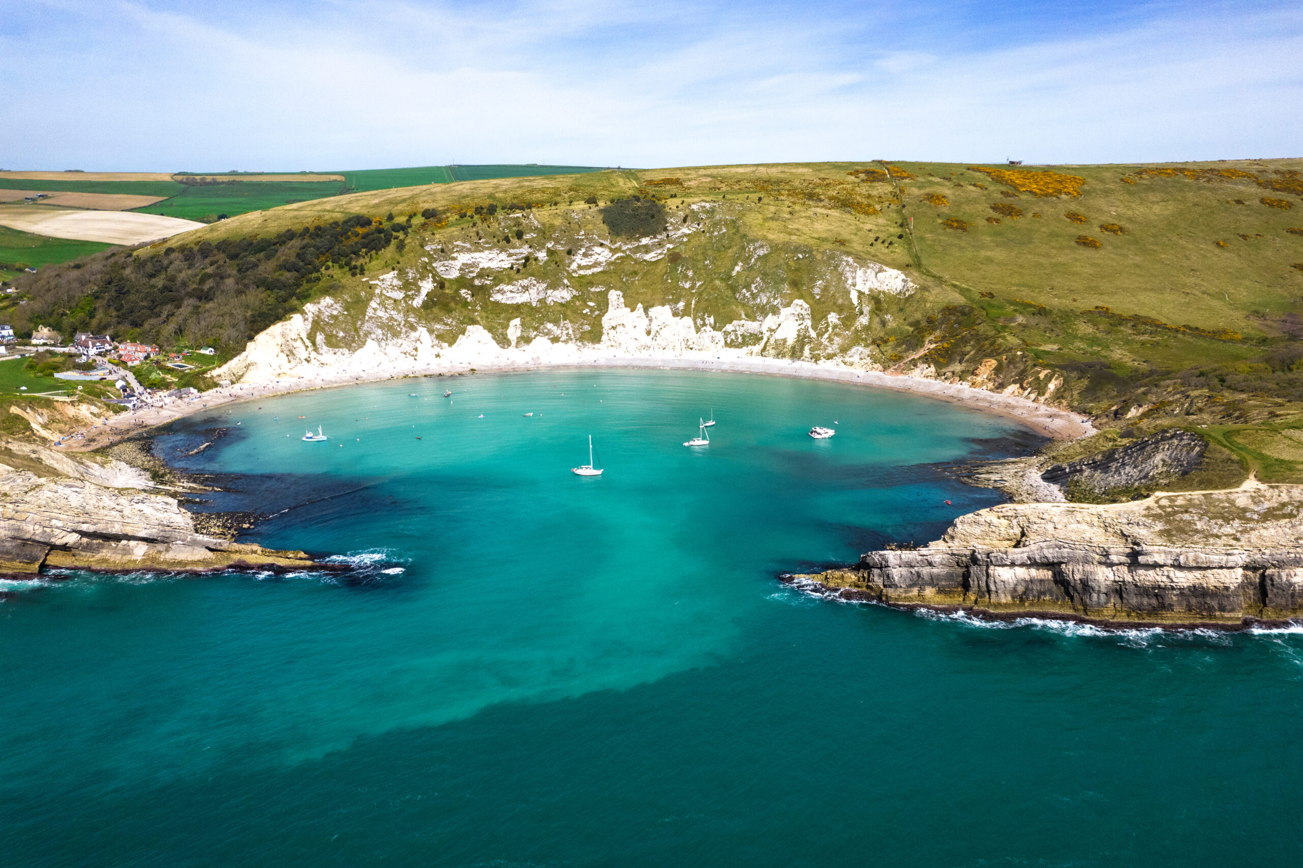 Aerial view of Lulworth Cove from above the sea: a wide oval shaped cove with very bright blue water, surrounded by grassy cliffs.