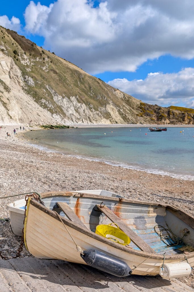 Small white wooden fishing boat on a white shingle beach with grass topped chalk cliffs and a very bright blue bay behind.