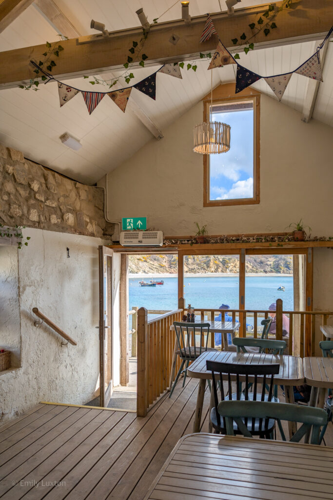 Interior of a small cafe with wooden tables and large bifold doors with a view of the sea. There is bunting hanging from the wooden beams on the cieling. 