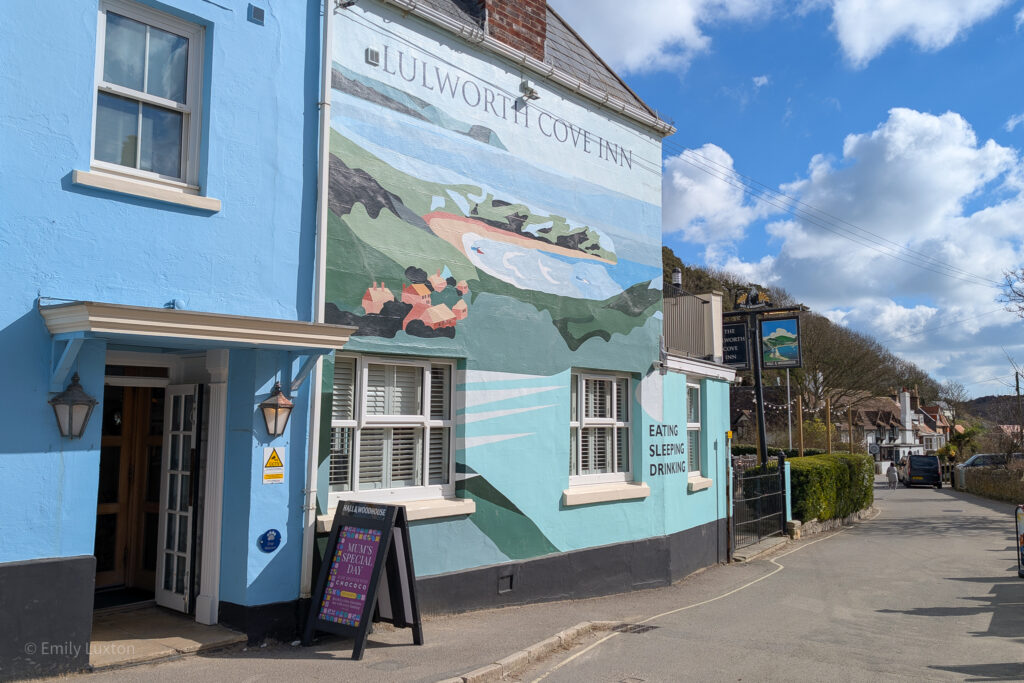 Exterior of a pub called Lulworth Cove Inn with a bright blue facade and a large mural of the cove and surrounding countryside. 