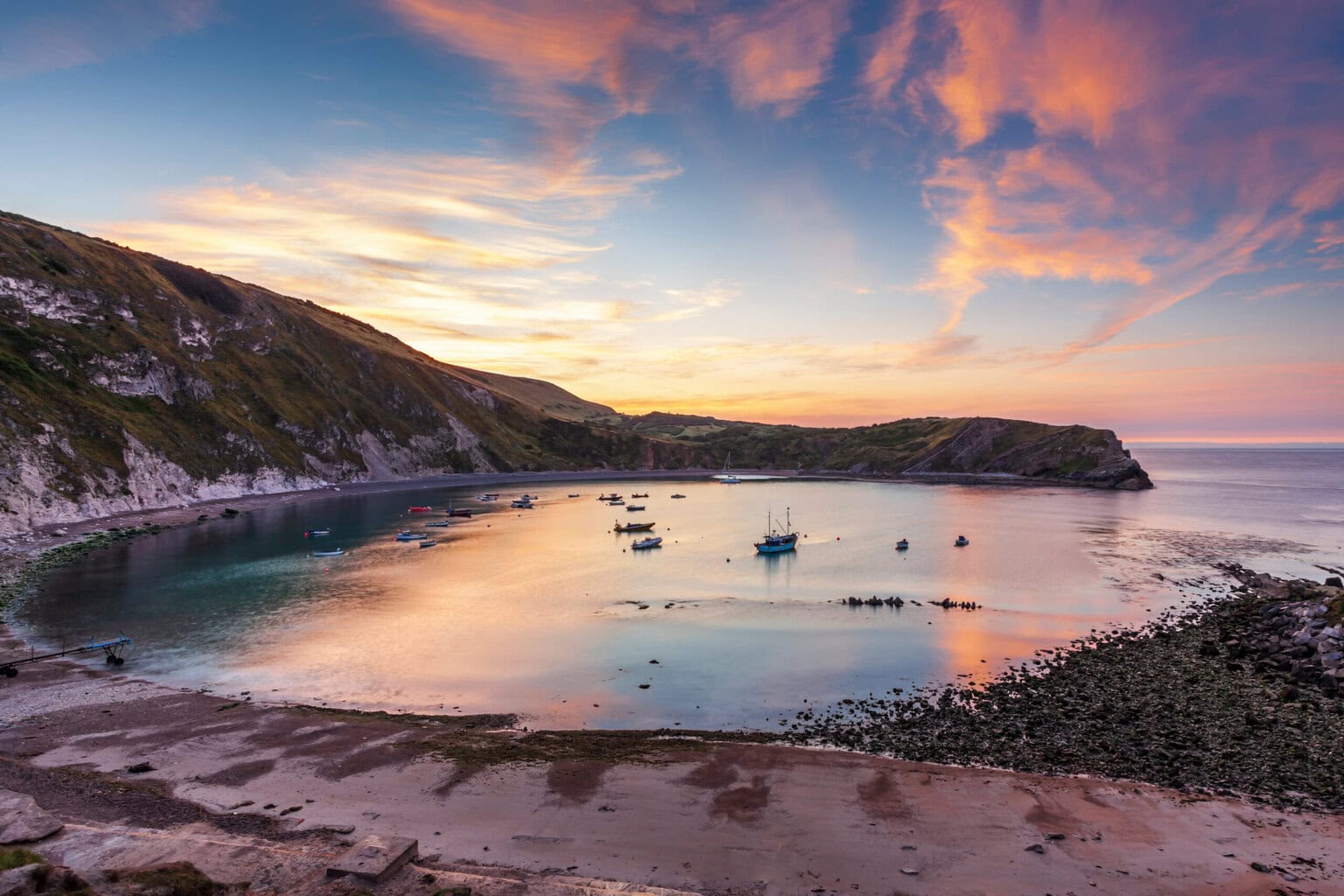 Lulworth Cove at sunrise with wjhite grassy cliffs to the left and pink sky reflected in a very circular bay of water where several small boats are moored.