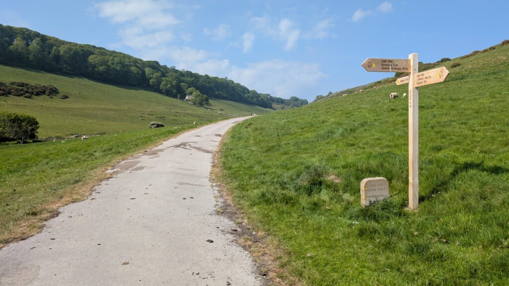 tarmac path leading throuhg a grassy valley with hills on either side and a wooden signpost
