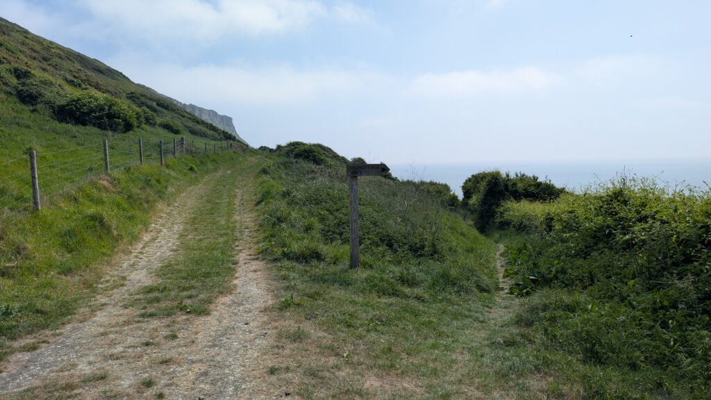 dirt path leading along the foot of a grassy sloping cliff with a wooden sign reading Chapman's Pool Beach pointing right