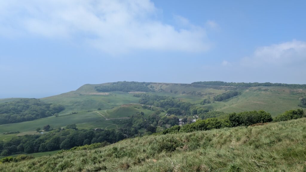 view of a wide green valley with a small woodland of green leafy trees and a large grey stone manor house just visible amidst the trees
