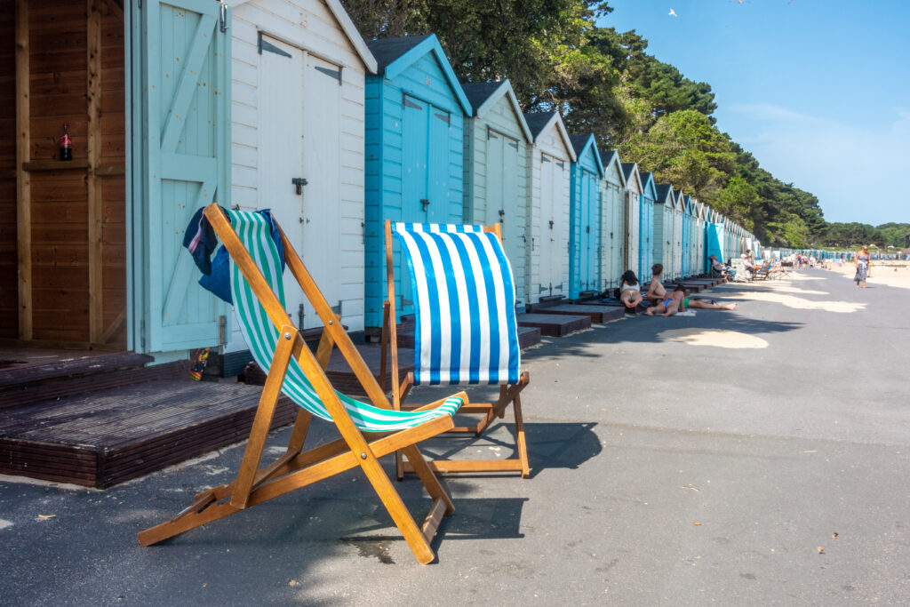 Blue deckchairs in front of pastel blue and white beach huts next to a sandy beach. 