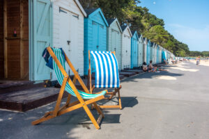 Blue deckchairs in front of pastel blue and white beach huts next to a sandy beach.