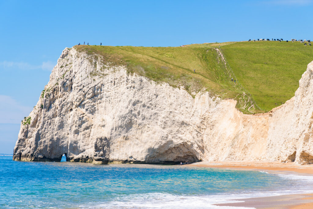 Large white chalk cliff jutting out into the sea with a small archway over the sea. There is a shingle beach in front. 