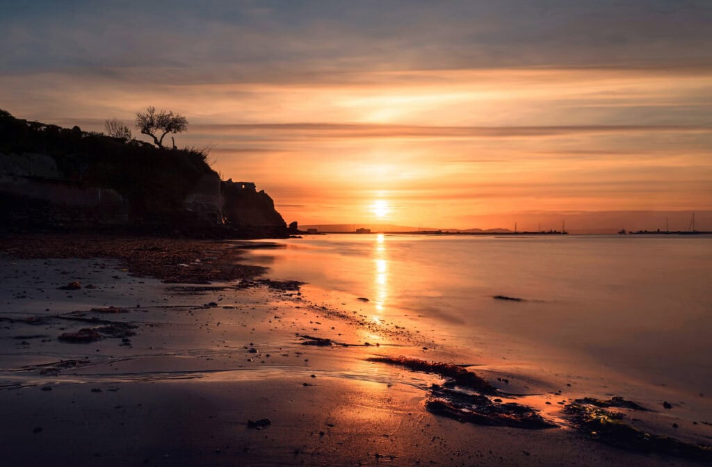 Sandy beach at sunset with low cliffs silhouetted against orange sky