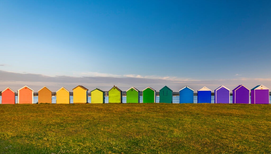 Rainbow of multi coloured beach huts at Hamworthy in Poole with a grassy field in the foreground and blue sea behind.