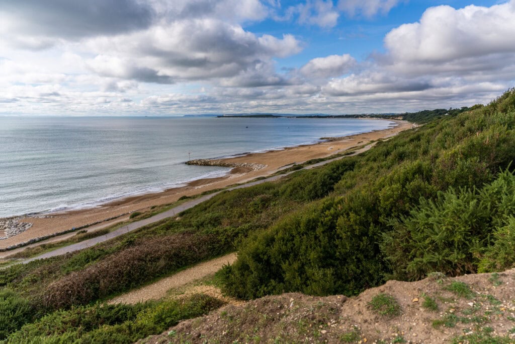 View from a grassy sloping cliffside of a shingle beach on a cloudy day with blue sea