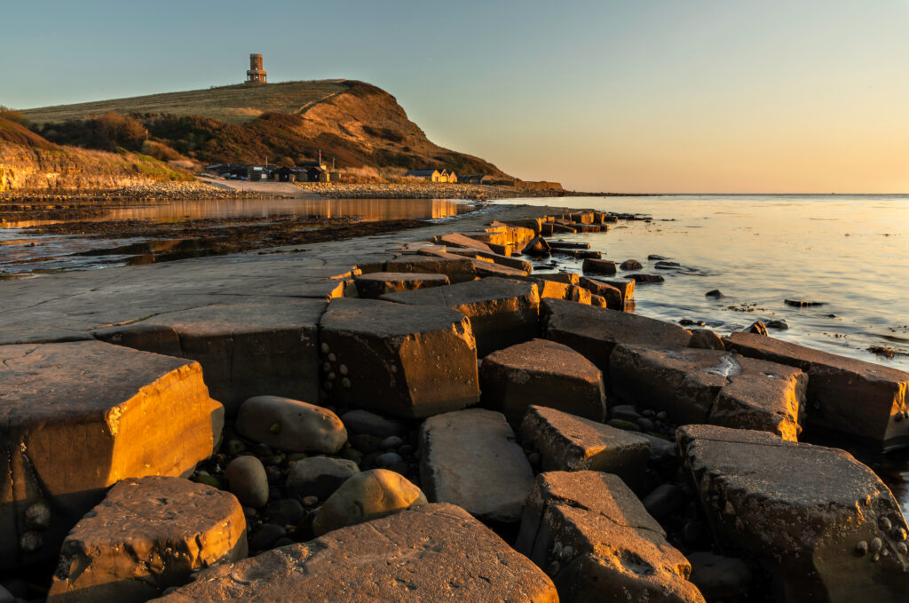 Juged grey rocky floor in the foreground with calm sea and a grassy headland with a stone tower in the distance