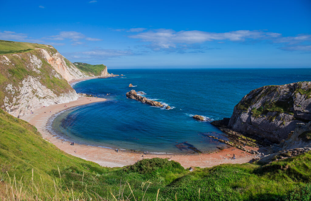 aerial view of a semi circle shaped cove with a white pebbly beach and chalk cliffs topped with grass