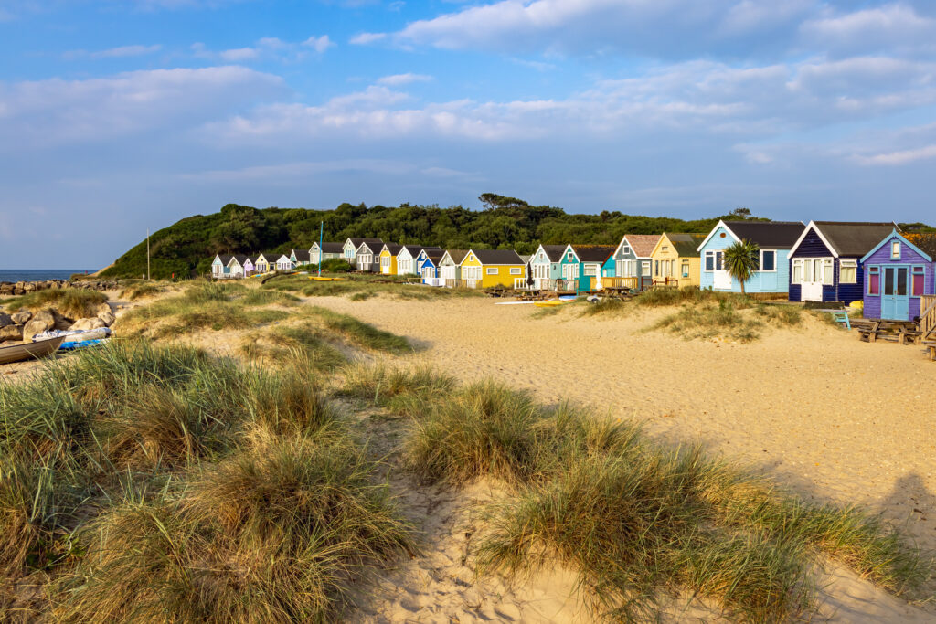 Row of wooden beach huts painted different pastel colours next to a white sand bewach with grassy dunes