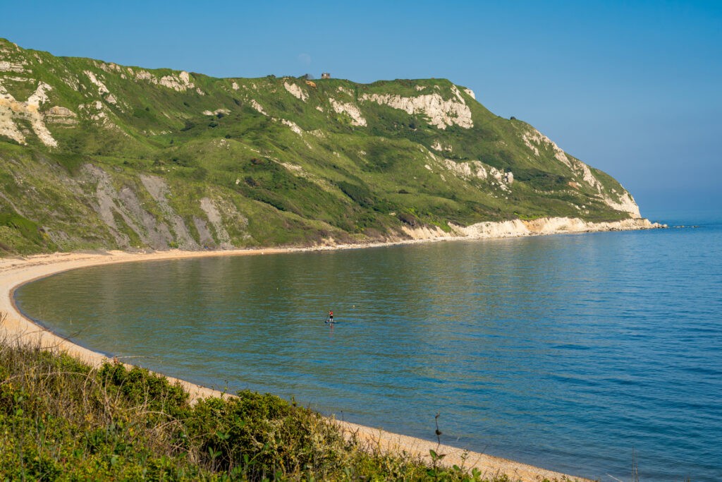 Large curved bay with very blue water and a shingle beach, there is a large grassy headland on the far side with chalky cliffs. 