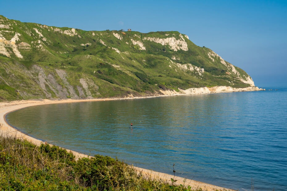 Large curved bay with very blue water and a shingle beach, there is a large grassy headland on the far side.