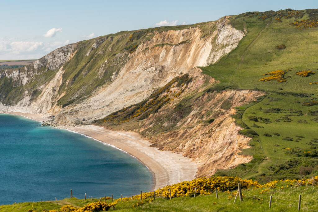 looking down at Worbarrow Bay from a grassy clifftop, the bay is very blue with a grey pebble beach backed by tall cliffs