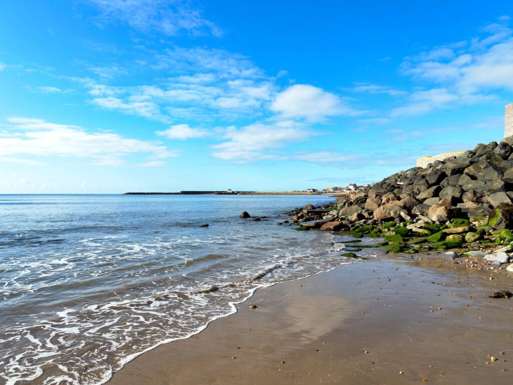 Small waves lapping at a sandy beach next to a large pile of grey rocks on a sunny day. 