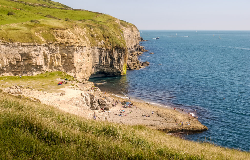 Grass topped cliffs with sheer grey rock faces and a lfat grey rock at the bottom near the sea where several people are standing