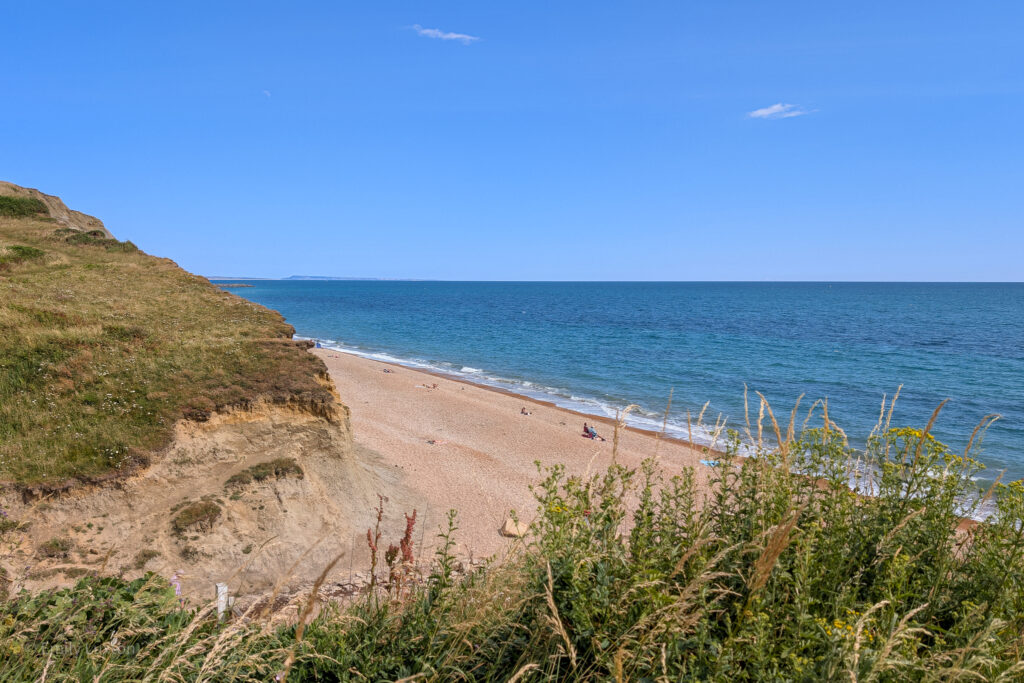 Looking down at a shingle beach and very blue sea from a grassy clifftop