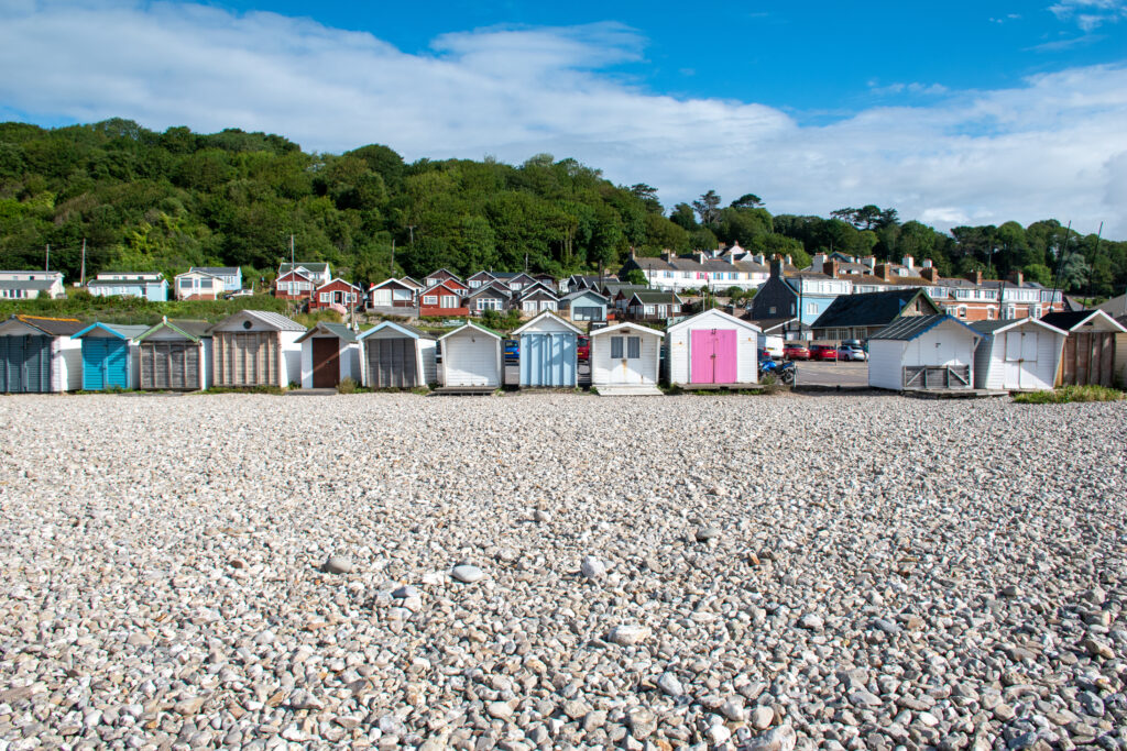White and grey pebble beach with a row of white woodne beach huts behind with different coloured doors. 