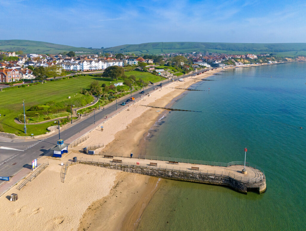 Aerial shot of a long sandy beach in Swanage with a stone jetty into the sea and a grassy hill behind the beach.