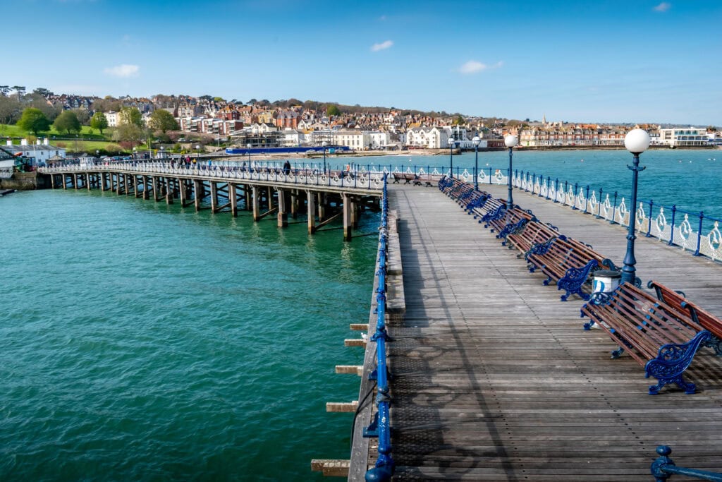 Looking back along a long wooden pier with  blue benches and sea on either side towards the town of Swanage