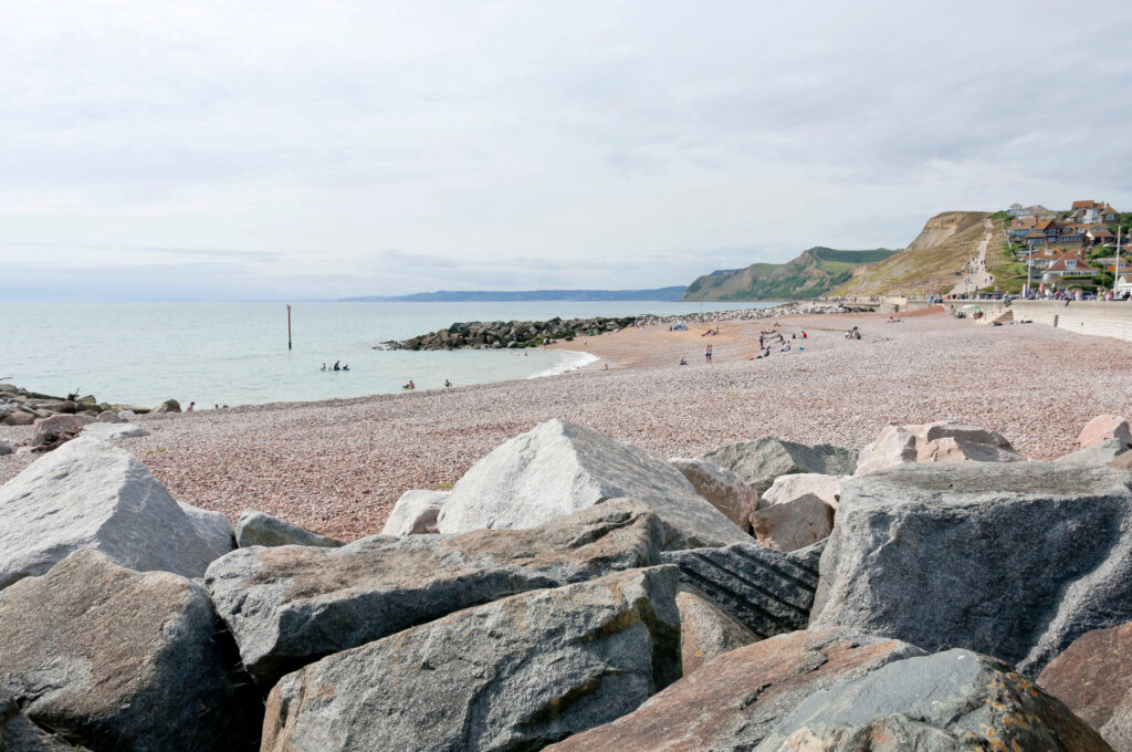 looking past grey rocks to a grey shingle beach on a cloudy day with a rock groyne and low grassy cliffs in the distance. 