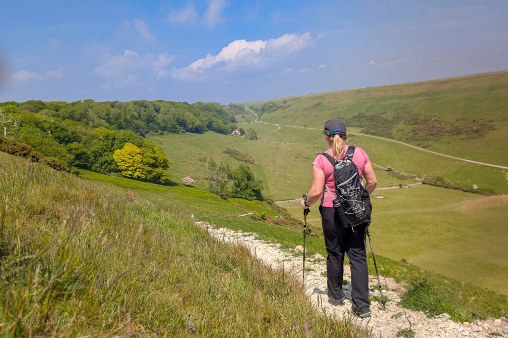 woman with hiking poles walking down a gravel path with a grassy valley beneath