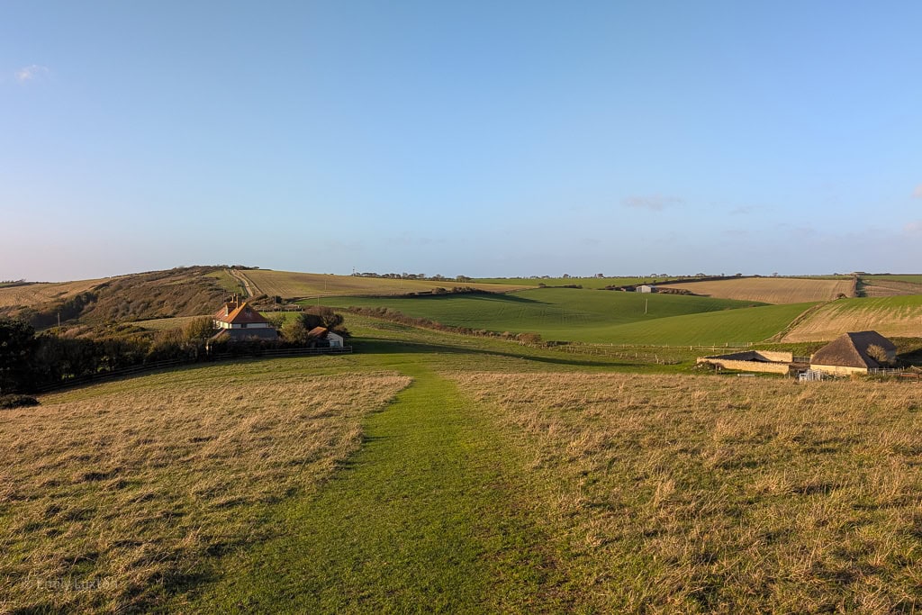 grassy track leading across a field towards a house with a hill behind